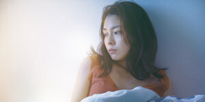 Eine Frau sitzt im Bett und lehnt an der Wand, die Bettdecke brusthoch gezogen, mit melancholischem Blick zur Seite