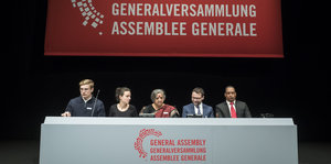 Fünf Menschen sitzen an einem Tisch, auf dem "Generalversammlung" steht