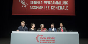 Fünf Menschen sitzen an einem Tisch, auf dem "Generalversammlung" steht