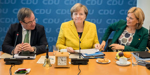 Armin Laschet, Angela Merkel und Julia Klöckner sitzen nebeneinander an einem Tisch, auf dem eine Uhr steht