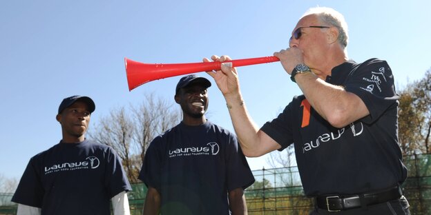 Franz Beckenbauer bläst in eine rote Vuvuzela. Zwei Männer beobachten ihn dabei