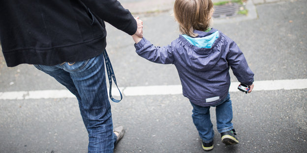 Ein Erwachsener hält ein Kind an der Hand, sie laufen eine Straße entlang