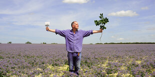 Bill Drummond steht in einem Feld und hält einen Zweig in der einen, einen Pinsel in der anderen Hand