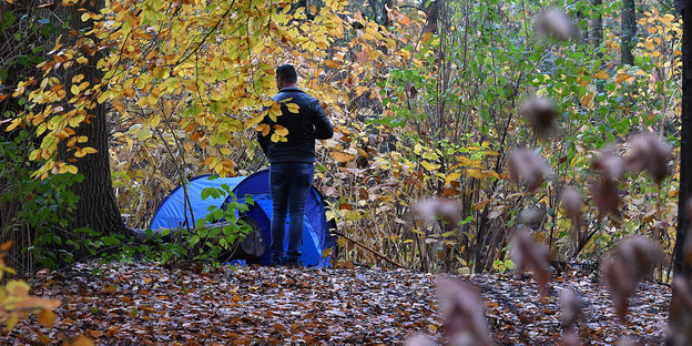 Ein Obdachloser steht zwischen Bäumen an einem Zelt.