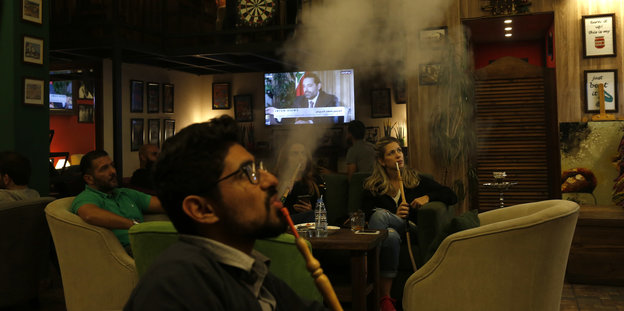 Menschen sitzen in einem Café und rauchen Wasserpfeife; hinter ihnen ist ein Fernseher zu sehen, auf dem die Ansprache des ehemaligen libanesischen Ministerpräsidenten Hariri läuft