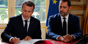 Emmanuel Macron und Christophe Castaner sitzen nebeneinander an eine Tisch. Macron unterschreibt ein Papier