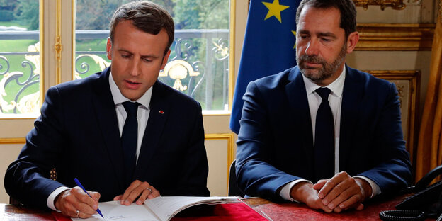 Emmanuel Macron und Christophe Castaner sitzen nebeneinander an eine Tisch. Macron unterschreibt ein Papier