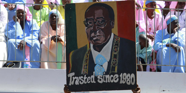 Hellblau und rosa gekleidete Menschen sind hinter einem Plakat mit dem Gesicht Mugabes versammelt, trusted since 1980 steht darauf