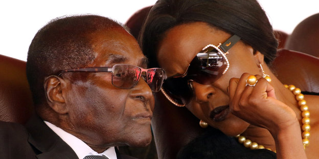 Mugabes Frau neigt sich zu ihrem Mann, der neben ihr sitzt und flüstert ihm etwas zu, beide tragen Sonnenbrillen