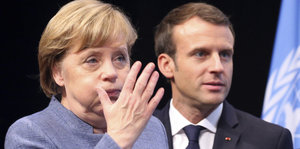 Angela Merkel bläst die Backen auf und hebt die Hand. Emmanuel Macron steht im Hintergrund