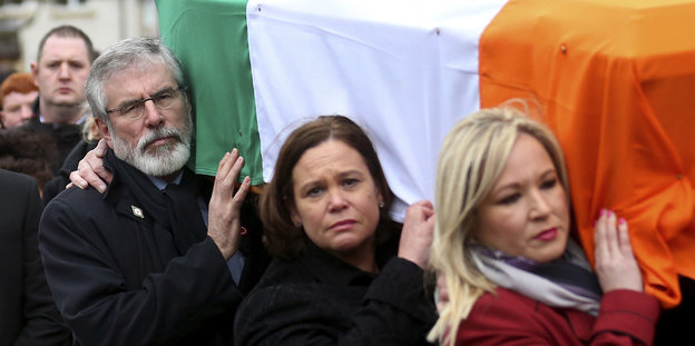 Drei menschen tragen einen Sarg, der mit einer irischen Flagge umwickelt ist