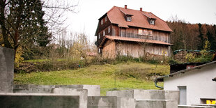 Betonstelen im Vordergrund, ein rustikales Holzhaus im HIntergrund