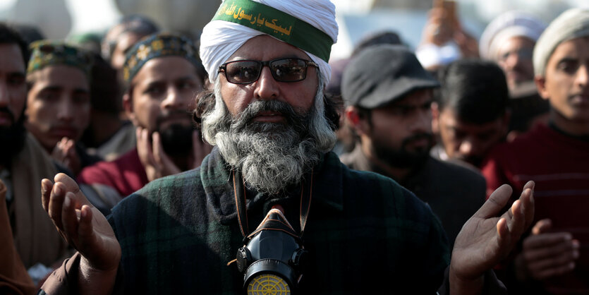 Ein Mann mit einem Turban, dem eine Gasmaske um den Hals hängt, hebt die Hände in die Höhe