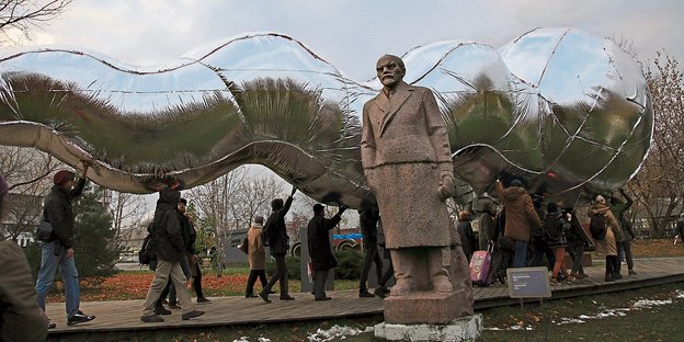 Menschen tragen eine riesige silberne Raupe über ihren Köpfen, im Vordergrund: eine Lenin-Statue