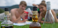 Im Vordergrund drei Flaschen Schnaps, im Hintergrund ein Junge und ein Mädchen, die mit Bechern anstoßen