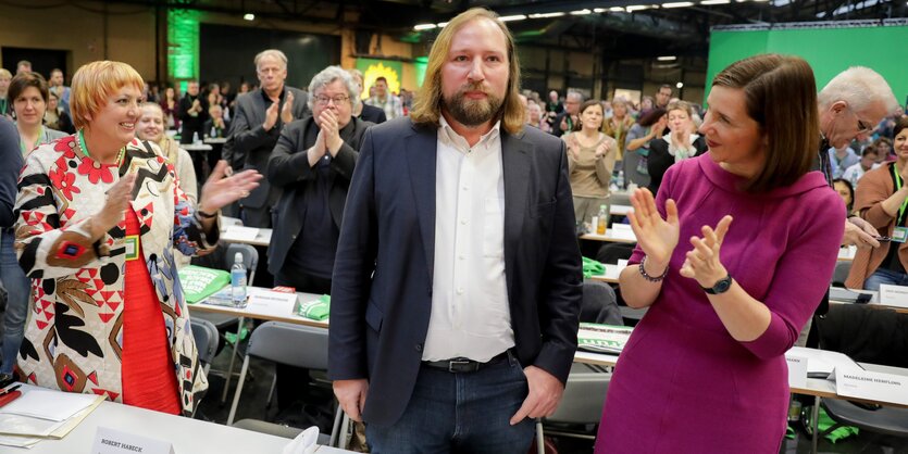 Ein Mann mit schulterlangen Haaren in der Mitte, zwei Frauen rechts und links von ihm, die applaudieren