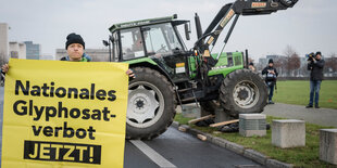 Ein Mann hält ein Schild, auf dem steht: Nationales Glyphosatverbot jetzt. Dahinter parkt ein Tecker.