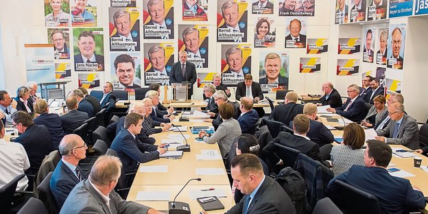 Blick in einen Raum mit vielen Männern, an den Wänden hängen Plakate von noch mehr Männern