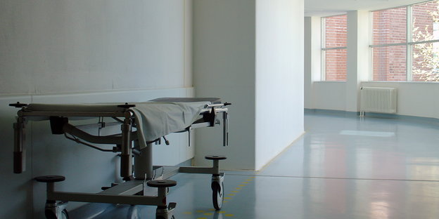 Eine Liege steht im weißen Krankenhausflur, im Hintergrund Fenster