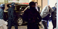 Ein Polizist mit Gewehr in der Hand steht vor mehreren Personen, die durch Bild laufen. Eine hat ihre Jacke über den Kopf gezogen