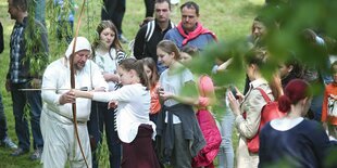 Ein kostümierter Mann leitet ein Mädchen im Bogenschießen an - hinter ihr eine Schlange weiterer Mädchen