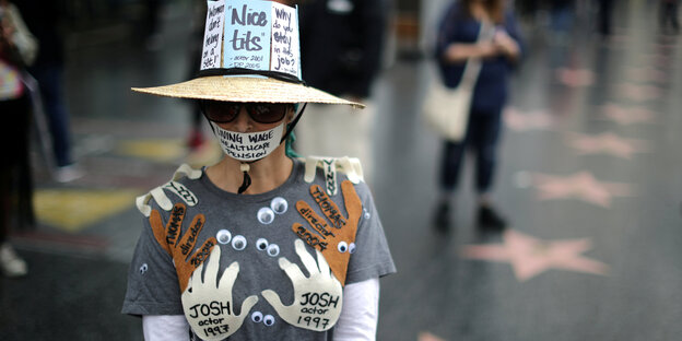 Eine Frau trägt auf ihrer Kleidung Papierstücke in Form von Händen, auf denen Namen und Jahreszahlen stehen, die sexuelle Übergriffe bezeichnen