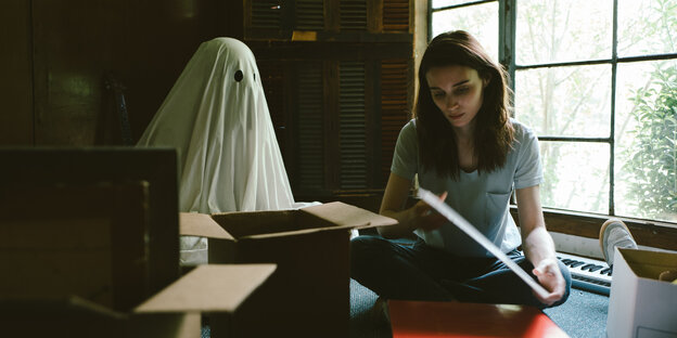 Eine Frau packt Bücher in einen Karton, daneben sitzt ein Gespenst