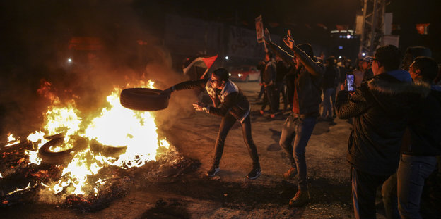 Jugendliche stehen um ein Feuer, einer wirft einen Autoreifen hinein, ein anderer filmt die Szene mit seinem Handy