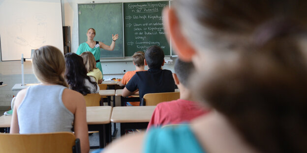 Eine Lehrerin steht an der Tafel und wendet sich ihrer Klasse zu