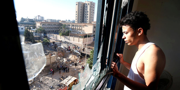 Mann im Unterhemd schaut sus dem Fenster auf Zerstörungen auf der Straße