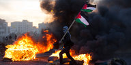 Ein Mann schwenkt die palästinensische Flagge, im Hintergrund brennen Reifen