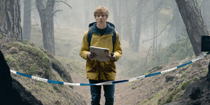 Ein junger Mann mit einer Taschenlampe und einer Landkarte steht alleine auf einem Waldpfad, der mit blau-weißem Flatterband abgesperrt ist