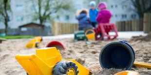 Ein Bagger und ein Eimer liegen im Sand, im Hintergrund sitzen Kinder