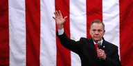 ein Mann mit erhobenem rechten Arm und einem Mikrofon in der Hand, im Hintergrund eine riesige US-Fahne