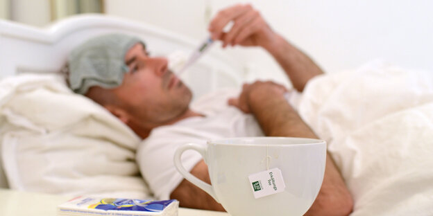 Ein Mann liegt mit Krankenhaushaube im Bett, ein Thermometer im Mund, eine Tasse Tee auf dem Nachttisch