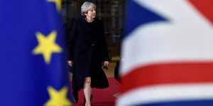 Eine Frau im schwarzen Mantel läuft auf einem roten Teppich, am Rand des Bilds sieht man eine europäische und eine britische Flagge