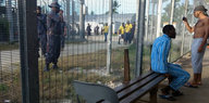 Zwei Menschen sitzen vor einem Zaun, dahinter befinden sich Polizisten