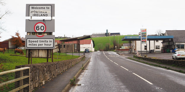 Eine Straße, die in ein Dorf an einem Hügel führt. Links ein Orts- und Geschwindigkeitsbegrenzungsschild