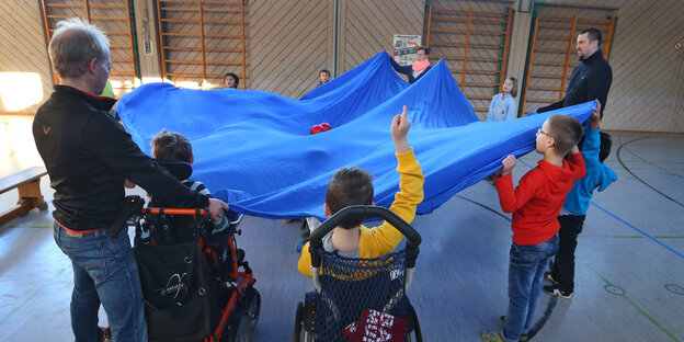 In einer Turnhalle heben Lehrer, gesunde Kinder und Kinder im Rollstuhl gemeinsam ein rundes Tuch in die Höhe.