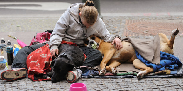 Eine junge Obdachlose sitzt mit gesenktem Kopf neben ihrem Hund auf dem Straßenpflaster.