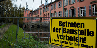 Ein langgestrecktes, rosafarbenes Haus, vor dem ein Bauzaun steht, an dem ein Schild „Betreten der Baustelle verboten! Eltern haften für ihre Kinder“ hängt.