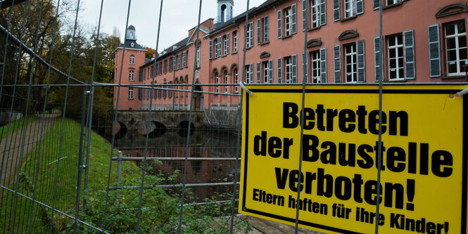 Ein langgestrecktes, rosafarbenes Haus, vor dem ein Bauzaun steht, an dem ein Schild „Betreten der Baustelle verboten! Eltern haften für ihre Kinder“ hängt.