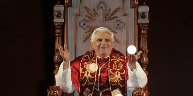 Ex-Papst Benedixt XVI. sitzend, die Hände in die Höhe haltend