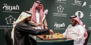 Zwei Männer mit Tüchern auf dem Kopf spielen lachend Schach, ein weiterer steht vor ihrem Tisch