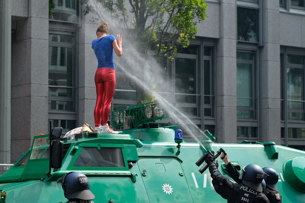 Auf einem grünen Polizeiräumfahrzeug steht eine Frau, Polizisten bespritzen sie mit Tränengas