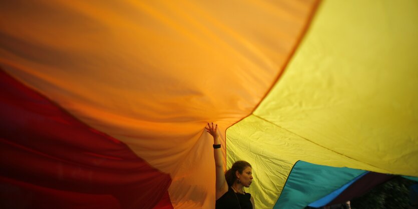 Eine Frau steht unter einer großen Regenbogenfahne und stützt sie mit ihrem Arm