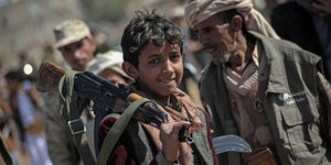 Ein Junge aus dem Jemen mit einem Gewehr über der Schulter. Hinter ihm stehen weitere Kämpfer der Huthi-Rebellen.