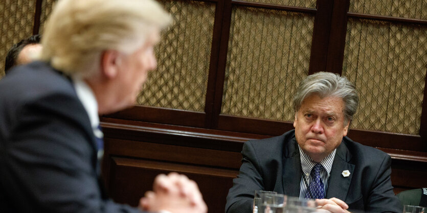 Donald Trumo sitzt an einem Tisch und beugt sich zu Steve Bannon