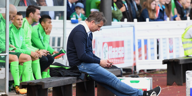 Ein mittelalter Mann sitzt auf einer Bank vor Fußball-Spielern und Fans und schaut auf sein Handy.