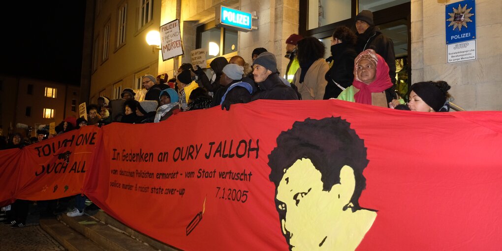 Viele Demonstrant*innen halten ein langes Transparent vor einem Gebäude stehend. Es ist Abend